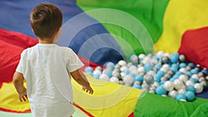 little Caucasian boy playing with a colorful piece of material and many balls on it, full shot kindergarten
