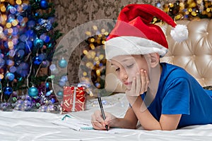 A little Caucasian boy in red Santa hat lies on the bed and writing a letter to Santa at home decorated for Christmas
