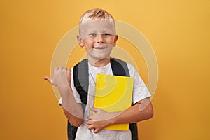 Little caucasian boy wearing student backpack and holding book pointing thumb up to the side smiling happy with open mouth