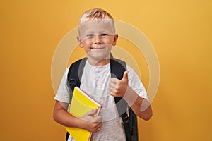 Little caucasian boy wearing student backpack and holding book smiling happy and positive, thumb up doing excellent and approval