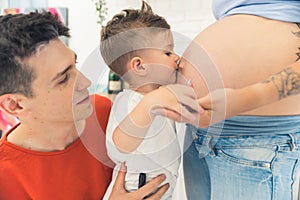 Little caucasian child son supported by his young adult father giving a kiss to his mother& x27;s pregnant belly