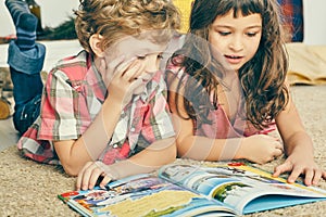 Little Caucasian curly boy and girl lying on the floor and reading an illustrated book.