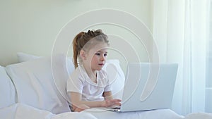 Little caucasian girl checks a laptop in bed. Modern Kids and Gadgets. The girl scrolls social networks. Electronic