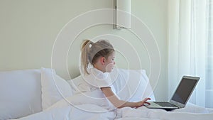 Little caucasian girl checks a laptop in bed. Modern Kids and Gadgets. The girl scrolls social networks. Electronic