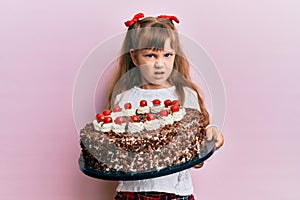 Little caucasian girl kid celebrating birthday holding big chocolate cake clueless and confused expression