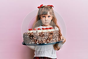 Little caucasian girl kid celebrating birthday holding big chocolate cake relaxed with serious expression on face