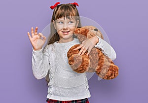 Little caucasian girl kid hugging teddy bear stuffed animal doing ok sign with fingers, smiling friendly gesturing excellent
