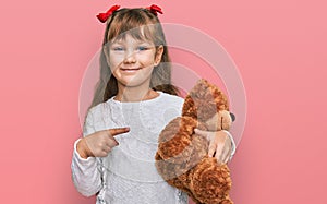 Little caucasian girl kid hugging teddy bear stuffed animal smiling happy pointing with hand and finger