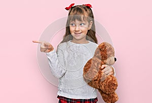 Little caucasian girl kid hugging teddy bear stuffed animal smiling happy pointing with hand and finger to the side