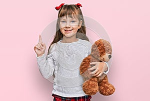 Little caucasian girl kid hugging teddy bear stuffed animal surprised with an idea or question pointing finger with happy face,