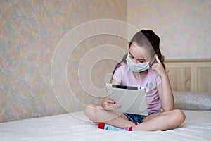 A little caucasian girl in a protective mask with a tablet sits on the bed in self-isolation mode at home