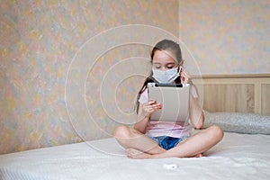 A little caucasian girl in a protective mask with a tablet sits on the bed in self-isolation mode at home