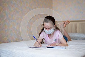 Little caucasian girl in a protective mask writes in a notebook during quarantine. Distance learning online education