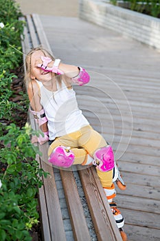 A little Caucasian girl in roller skates sits on a bench.