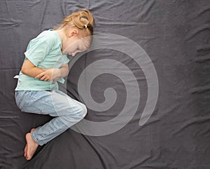 A little caucasian girl of seven years old lies on a gray bed with her legs crossed and holds her stomach with her hands