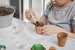 Little caucasian girl sits at a table outdoors in the backyard of the house and pours soil into a cardboard cup