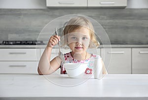 Little caucasian girl sitting at kitchen table with spoon and bowl, child has a meal, breackfast. Kid\'s nutrition concept