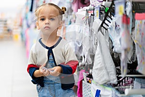 A little Caucasian girl is standing near a shop window with clothes and underwear in a large store. Little buyer