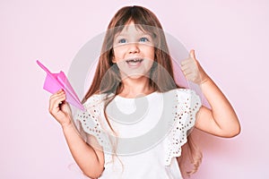 Little caucasian kid girl with long hair holding paper airplane smiling happy and positive, thumb up doing excellent and approval