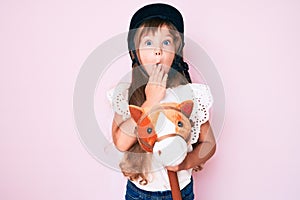 Little caucasian kid girl with long hair riding horse toy wearing vintage helmet covering mouth with hand, shocked and afraid for