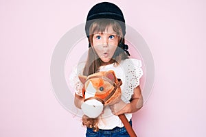 Little caucasian kid girl with long hair riding horse toy wearing vintage helmet scared and amazed with open mouth for surprise,