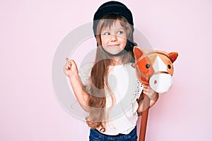 Little caucasian kid girl with long hair riding horse toy wearing vintage helmet smiling happy pointing with hand and finger to