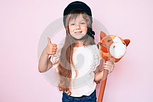 Little caucasian kid girl with long hair riding horse toy wearing vintage helmet smiling happy and positive, thumb up doing
