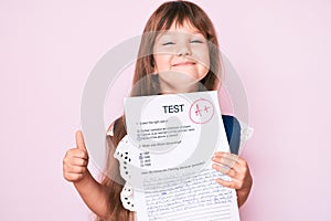 Little caucasian kid girl with long hair showing a passed exam from primary school smiling happy and positive, thumb up doing