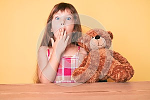 Little caucasian kid girl with long hair sitting on the table with teddy bear covering mouth with hand, shocked and afraid for