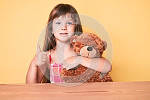 Little caucasian kid girl with long hair sitting on the table with teddy bear smiling happy and positive, thumb up doing excellent