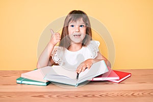 Little caucasian kid girl with long hair studying for school exam smiling happy and positive, thumb up doing excellent and