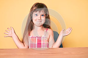 Little caucasian kid girl with long hair wearing casual clothes sitting on the table clueless and confused with open arms, no idea
