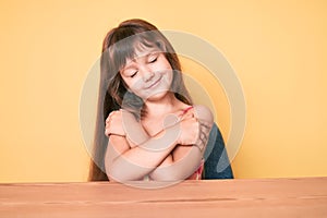 Little caucasian kid girl with long hair wearing casual clothes sitting on the table hugging oneself happy and positive, smiling