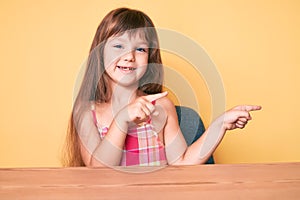 Little caucasian kid girl with long hair wearing casual clothes sitting on the table smiling and looking at the camera pointing
