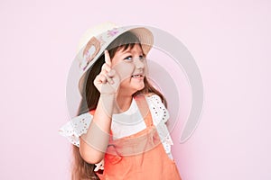 Little caucasian kid girl with long hair wearing spring hat smiling with an idea or question pointing finger up with happy face,