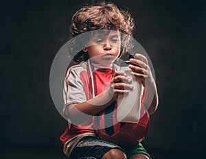 Little champion boy in sportswear holds a ball and drinking water from a bottle. Isolated on a dark textured background.