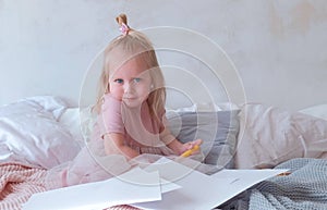 Little charming blond girl in pink dress holding a felt-pens and sitting on bed among paper.