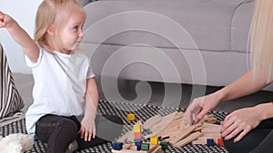 Little charming blond girl playing wood blocks with her mom sitting near the sofa.