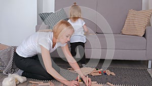 Little charming blond girl playing wood blocks with her mom sitting near the sofa. Build the tower and broke it.