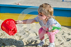Little charming girl baby playing on the playground in the sandbox sand mound in the bucket with a shovel and rake