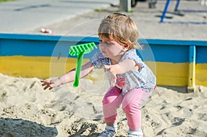 Little charming girl baby playing on the playground in the sandbox sand mound in the bucket with a shovel and rake