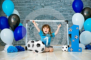 Little cheerful child dressed in sports clothes sitting on the floor near a football goal, looking at a big soccer ball