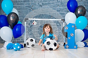 Little cheerful child dressed in sports clothes sitting on the floor near a football goal, looking at a big soccer ball. The first