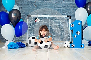 Little cheerful child dressed in sports clothes sitting on the floor near a football goal, looking at a big soccer ball. The first