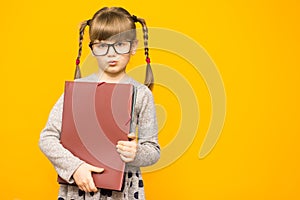 Little cheerful girl in glasses with serious face and funny pigtails holding school books against yellow