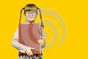 Little cheerful girl in glasses with serious face and funny pigtails holding school books against yellow