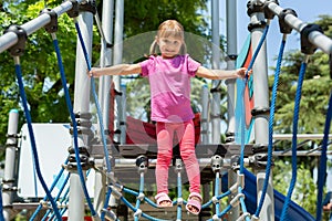 A little cheerful girl walks on a rope bridge on a playground in the park.