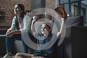 Little cheering boy with baseball mitt and ball sitting in living room with his mother and watching a game