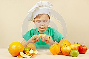 Little chef peeling fresh orange at table with fruits