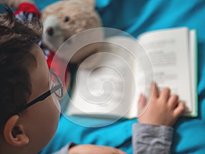 Little child, 8 years old boy reading a book at home with his toy teddy bear